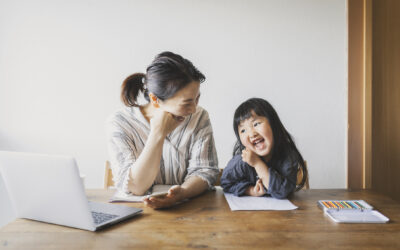 AAPI mom on laptop and daughter coloring are laughing together at the table