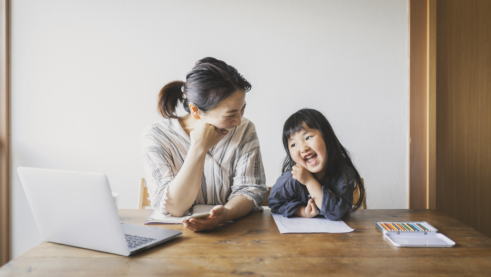 AAPI mom on laptop and daughter coloring are laughing together at the table